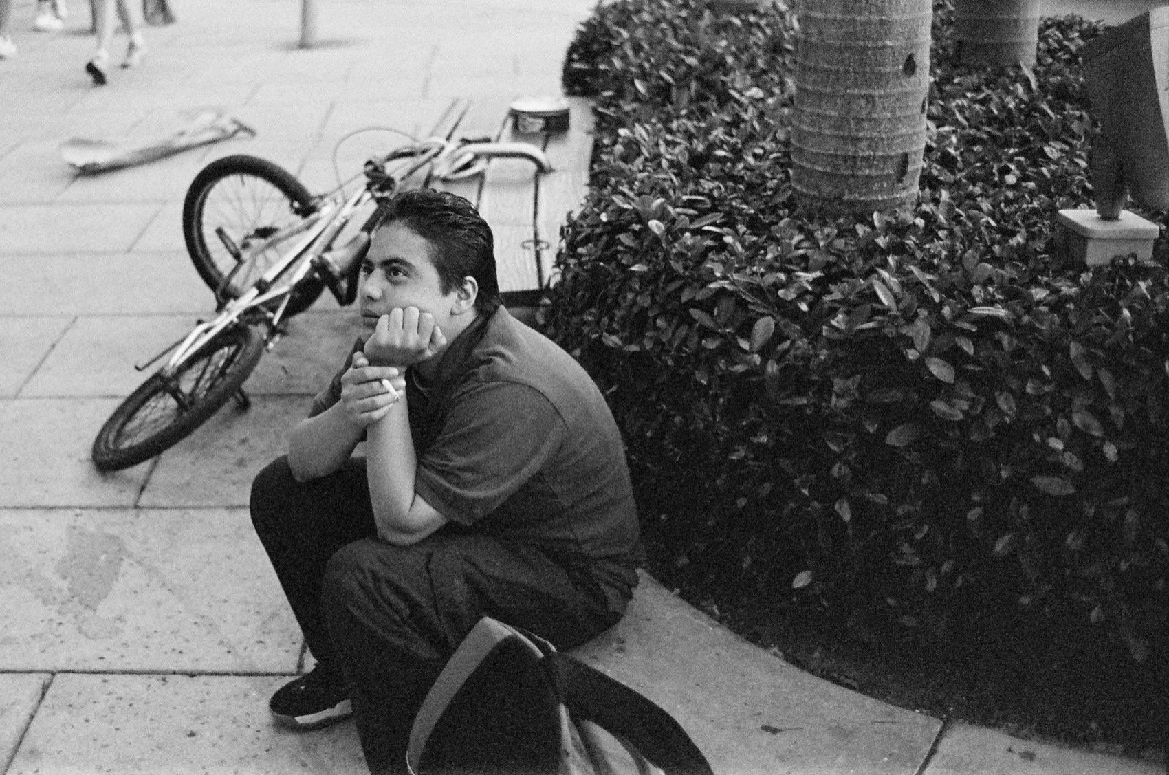 A boy crouched on a kerb beside a dropped BMX, chin resting on his hands.