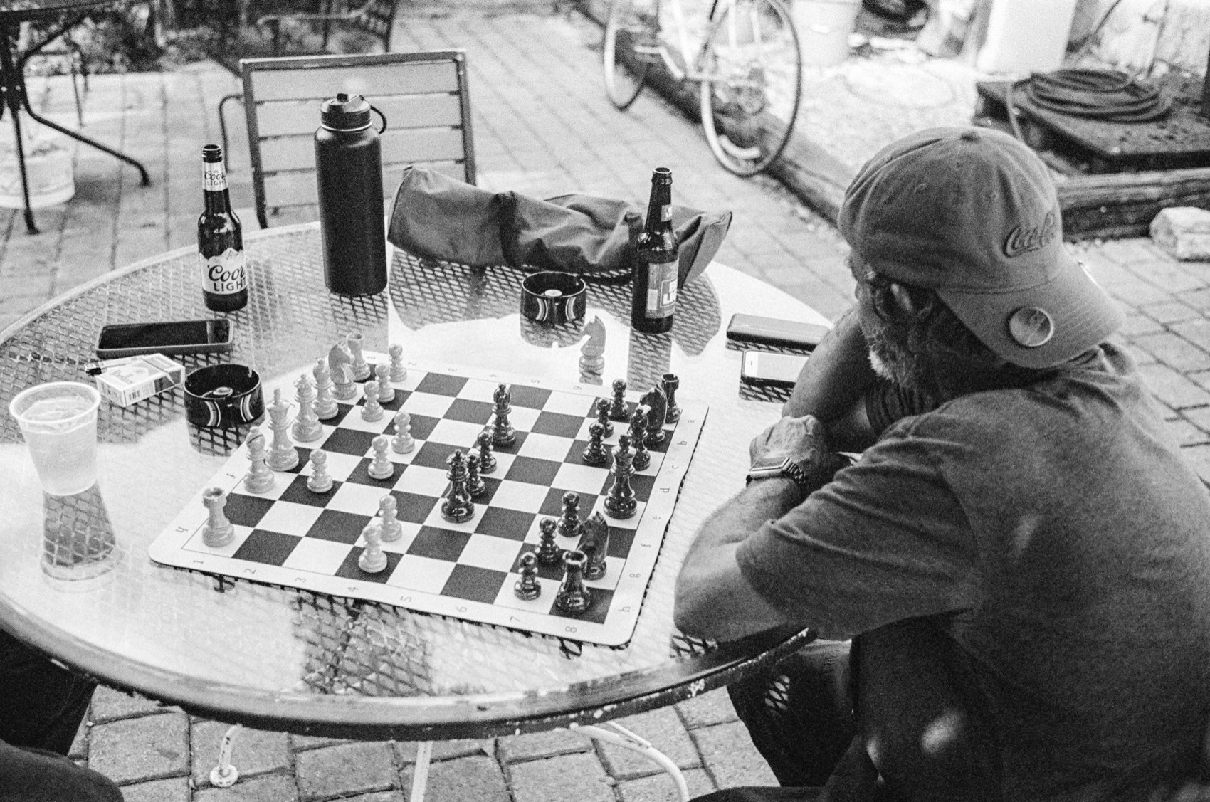 A chess game part-played on an outdoor table, bottles and glasses around it.