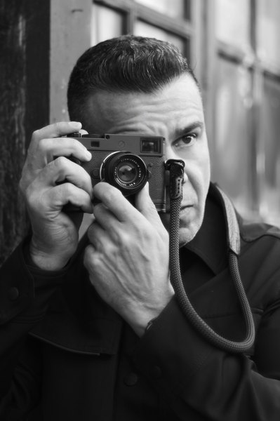 Gaston Shutters raising a Leica rangefinder to his eye, photographed in black and white against a window