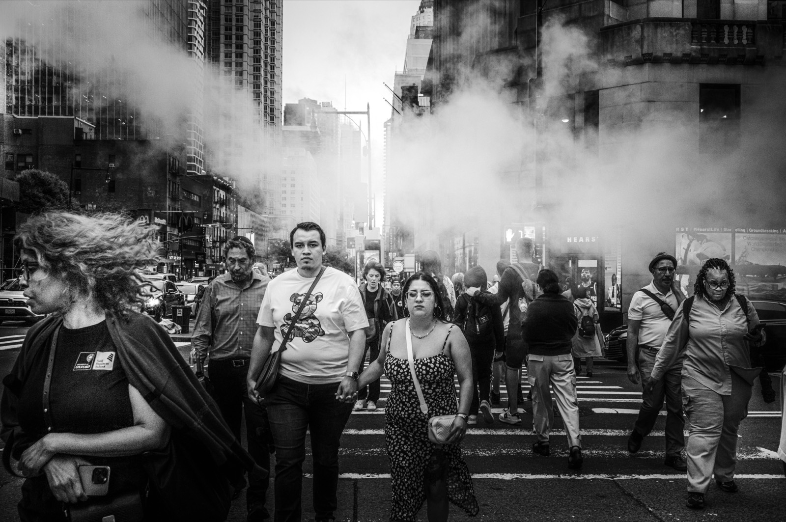 A crowd crossing an avenue in front of a large plume of backlit street steam.