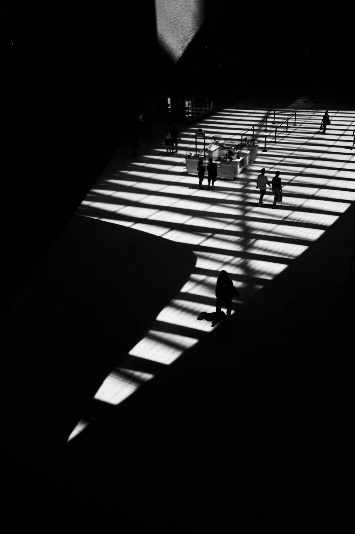 A diagonal shaft of daylight across a dark plaza, with figures reduced to specks inside it.