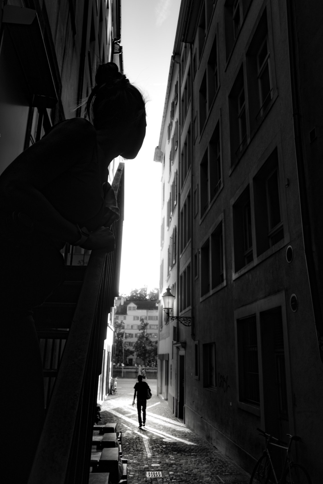A woman leaning over a railing above a narrow Zurich alley, a man walking away down the cobbles into the light.
