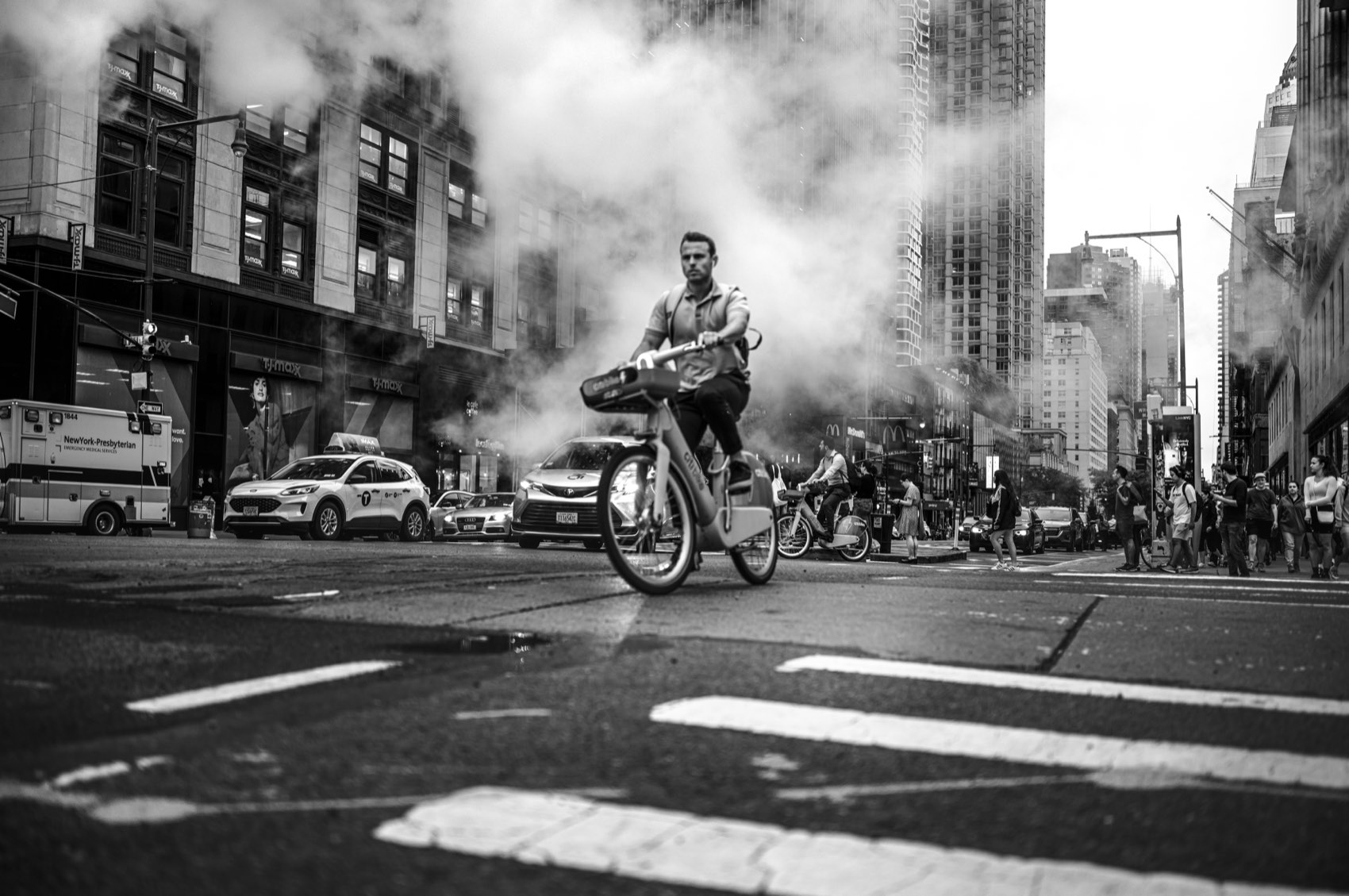 A cyclist crossing an avenue in front of a large plume of backlit street steam.