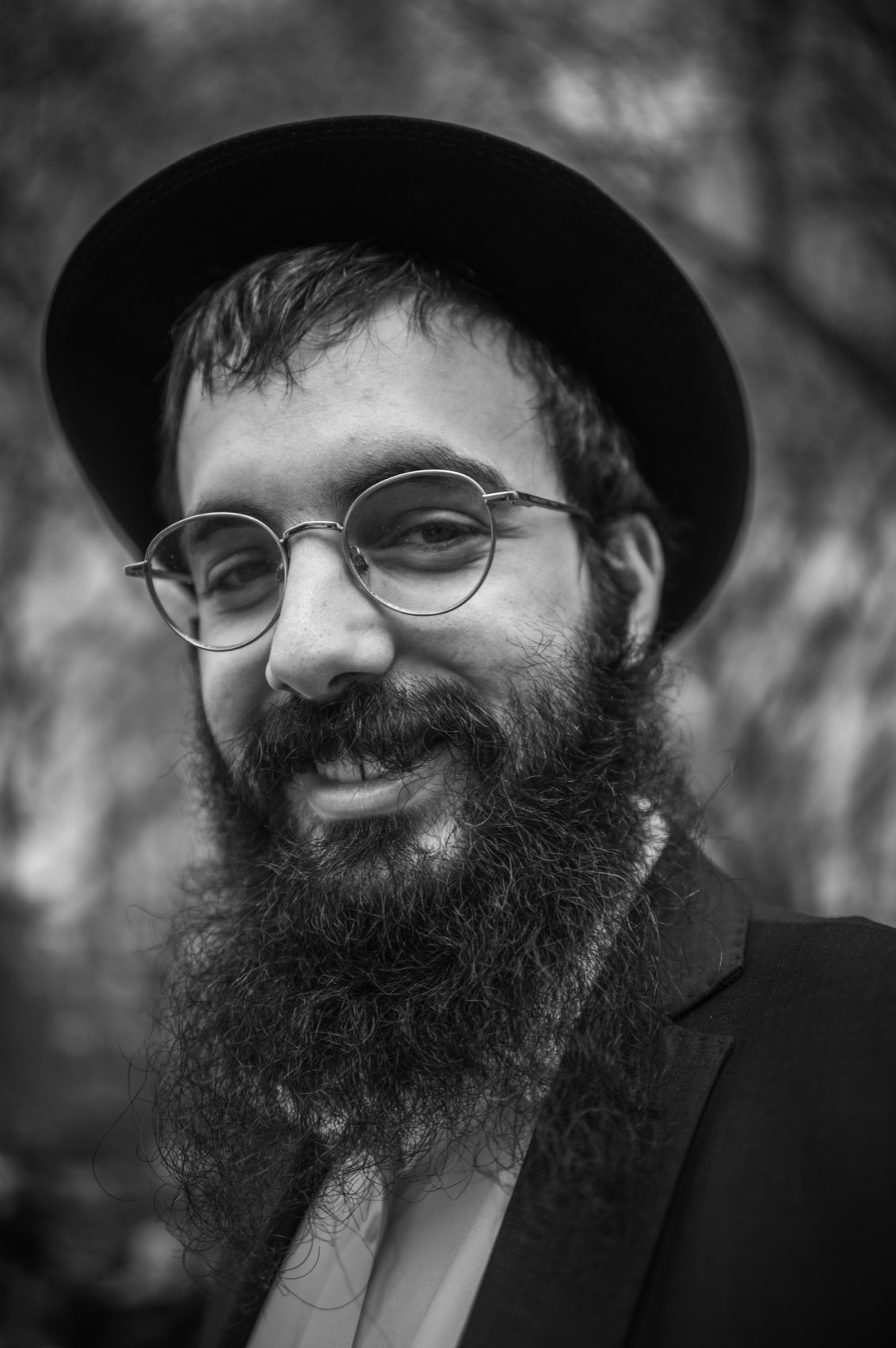 A bearded man in a black hat and round glasses, photographed close and looking straight into the lens.